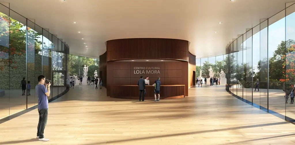 Fotografía del interior del Centro Cultural Lola Mora, mostrando un amplio pasillo con suelos de madera, paredes de vidrio que permiten la entrada de luz natural, y una recepción central de madera curvada. Al fondo, se pueden apreciar visitantes admirando esculturas en exhibición.